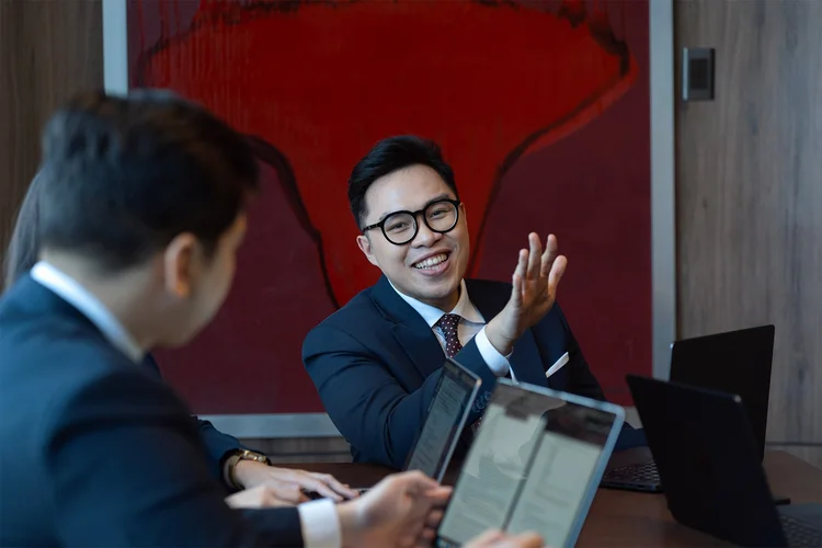 Two men in business suits discussing with laptops open at a meeting table, one smiling and gesturing.