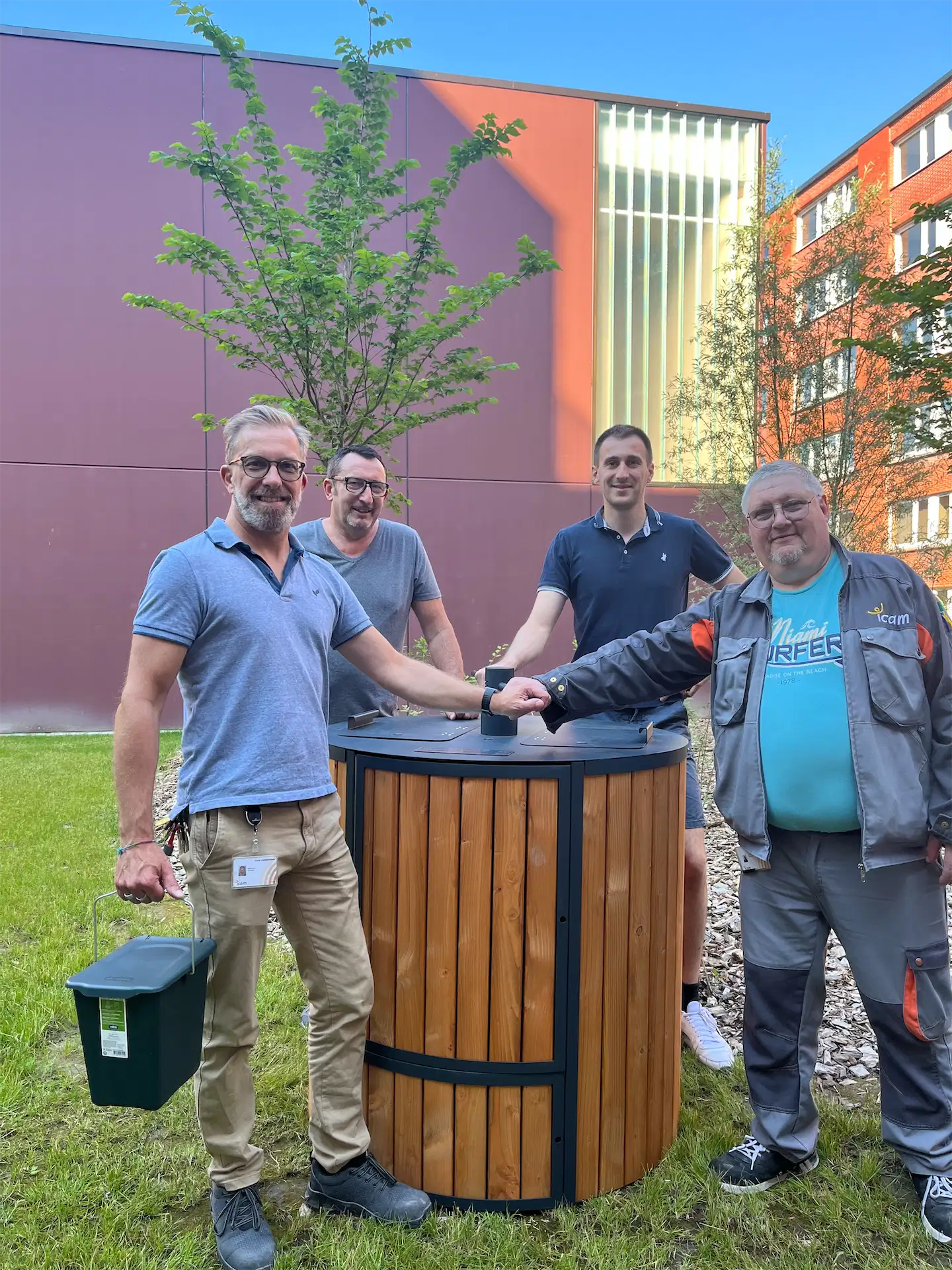 Four men standing outdoors around a wooden cylindrical bin, two of them fist bumping and one holding a small green container. Easy To Compost