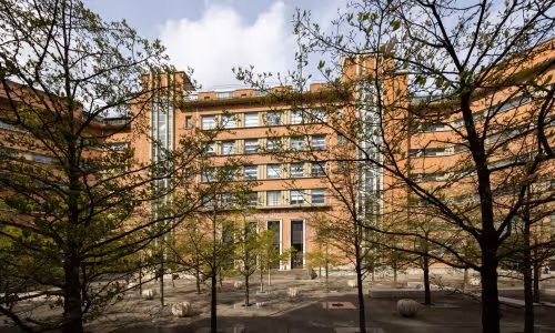 Five-story brick building with tall vertical windows surrounding a courtyard with young trees and stone benches.