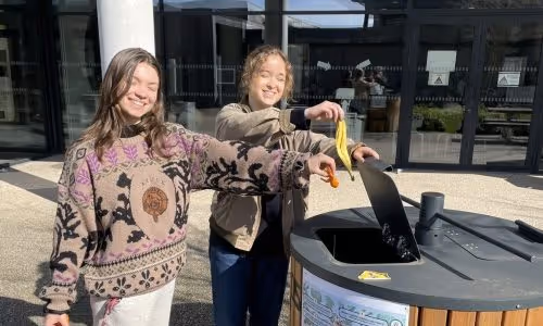 Two smiling women outdoors throwing food waste into a compost bin.