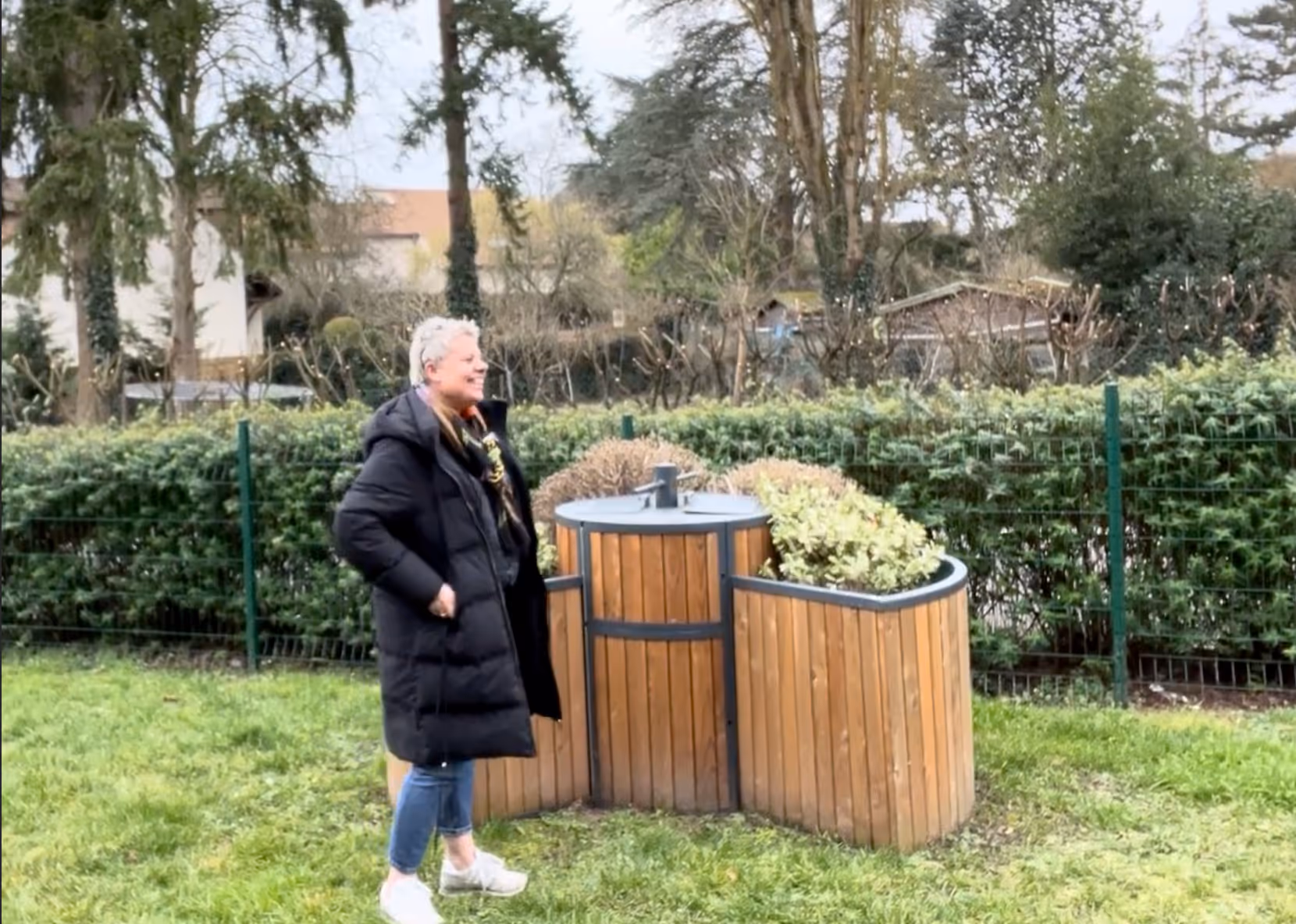 Woman in black coat stands on grass near a wooden planter with green and brown plants in a fenced garden. ABC Composter - Easy To Compost.