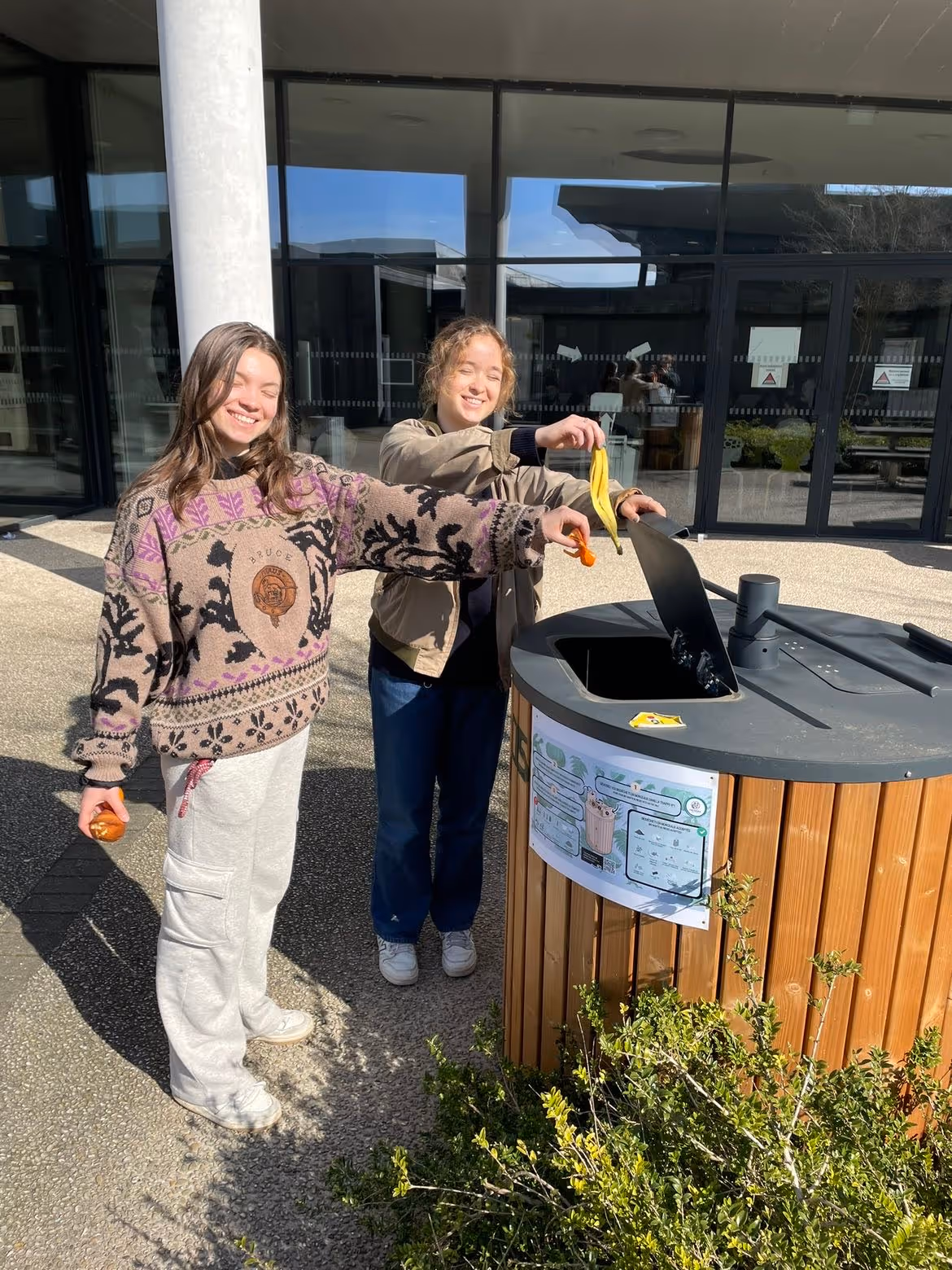 Two smiling young women outdoors, disposing of food waste into a round wooden compost bin. ABC Composter - Easy To Compost.