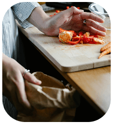 Person scraping food scraps from a cutting board into a brown paper bag.