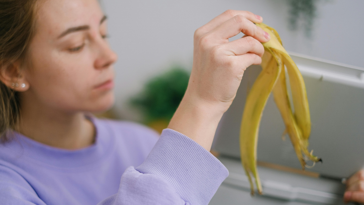 Person in a lavender sweatshirt holding banana peels over an open trash bin.