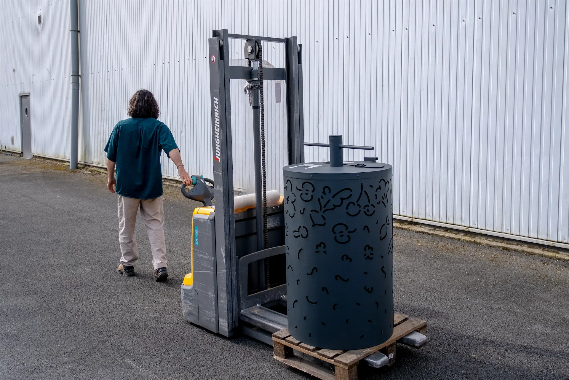 Livraison d'un composteur. Person operating a forklift carrying a composter with cut-out patterns on a wooden pallet outside a corrugated metal building.