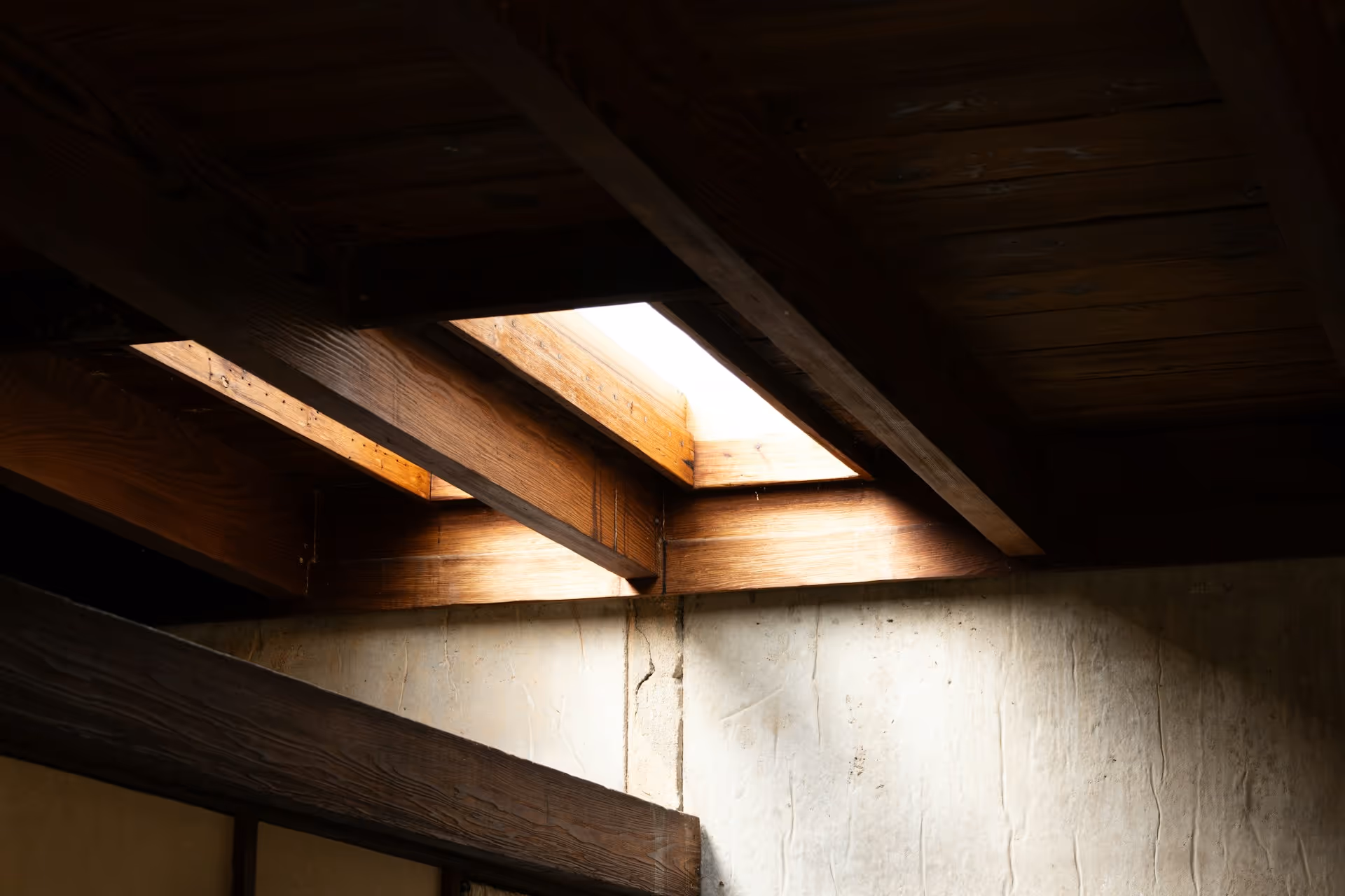 Sunlight streaming through rectangular skylights framed by wooden beams onto a textured concrete wall.