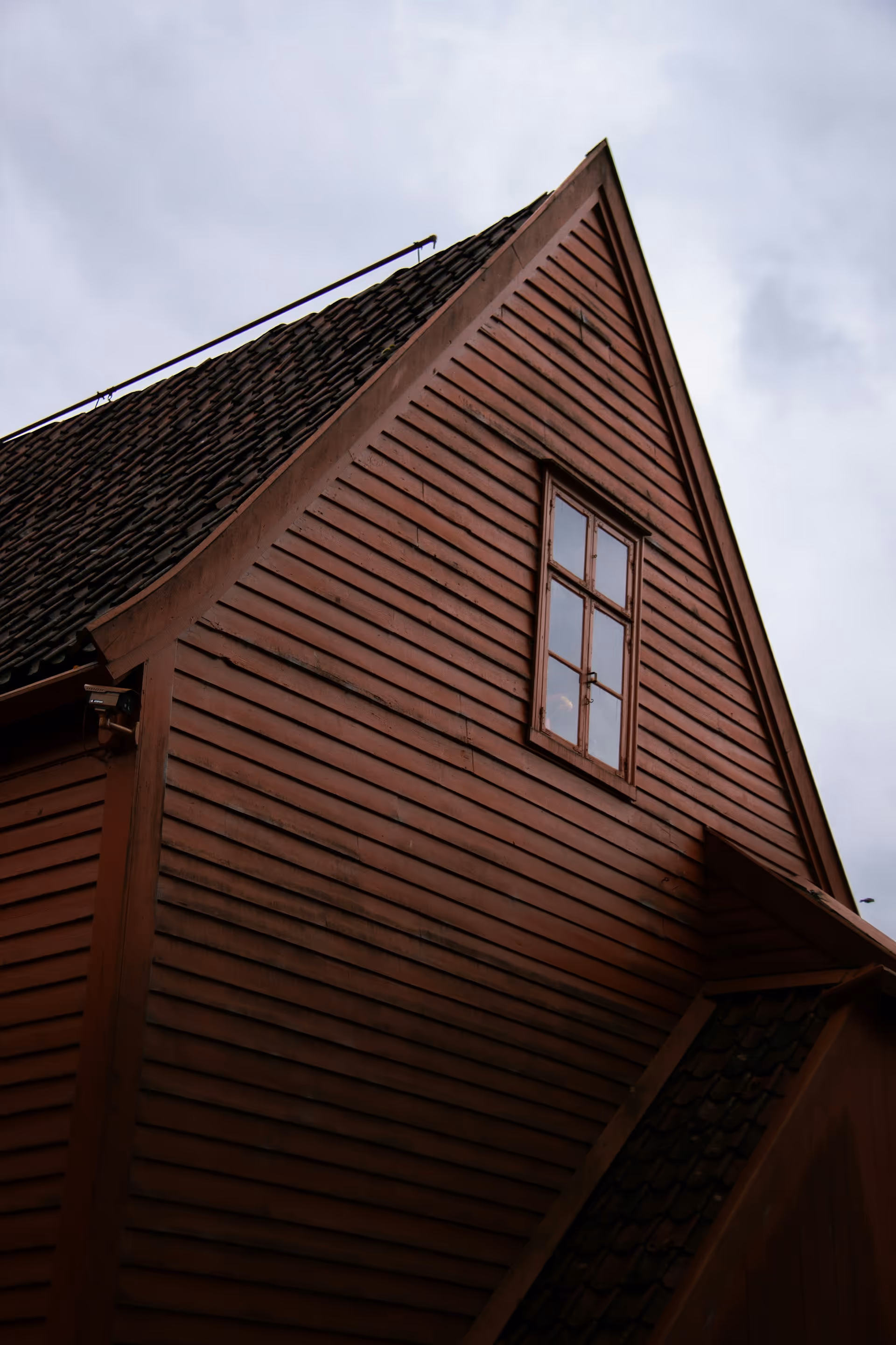Close-up of the upper side of a weathered red wooden house with a tiled roof and a window under a cloudy sky.