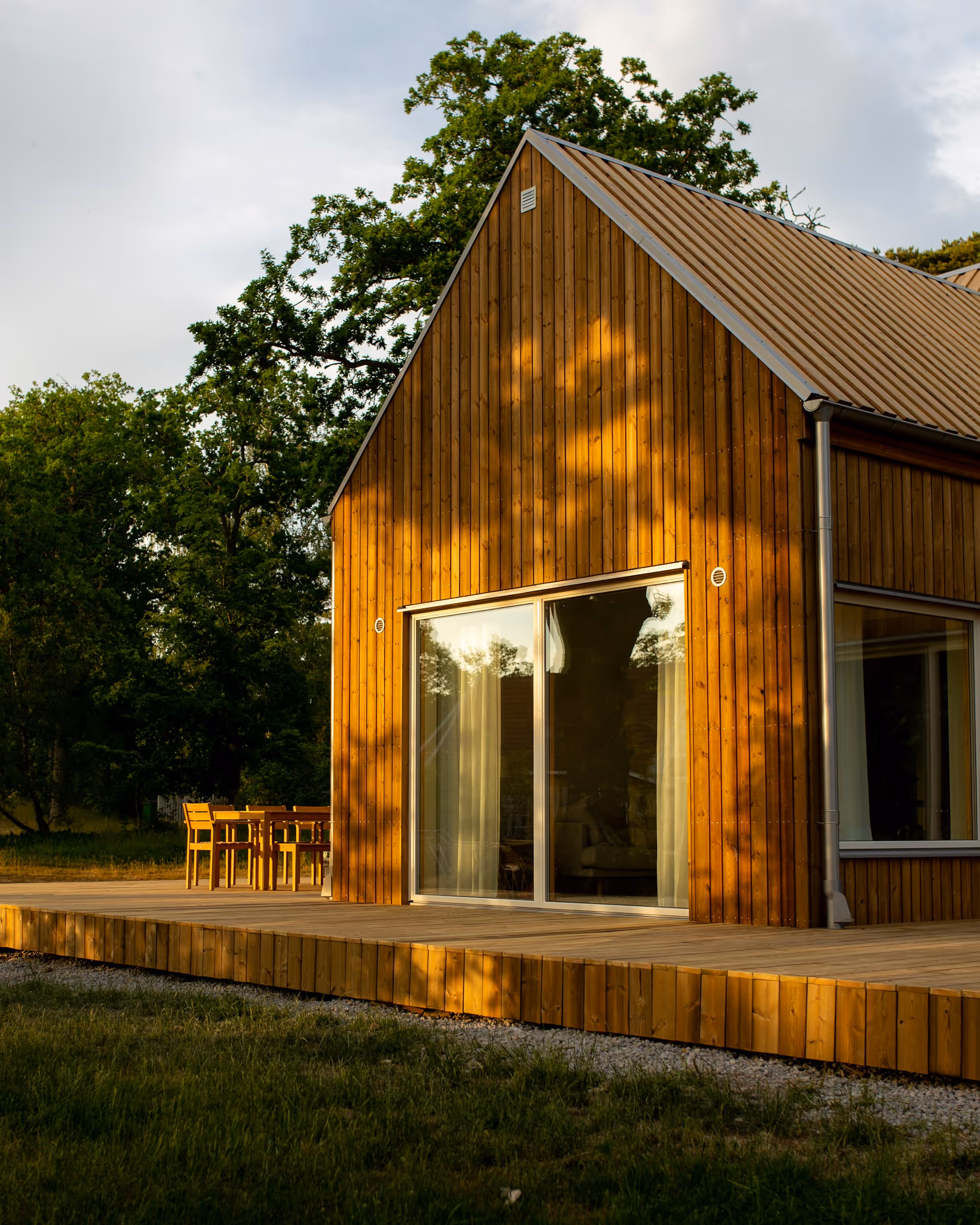 Modern wooden house with large sliding glass doors and a deck with outdoor furniture, surrounded by trees in the late afternoon light.