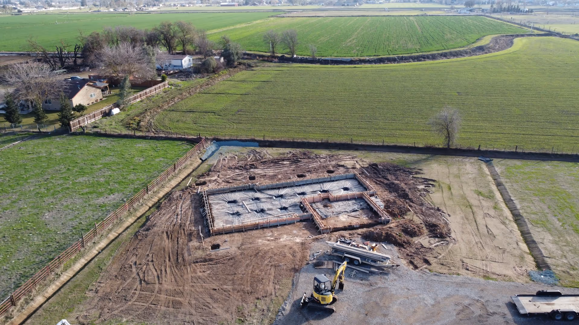 Aerial view of a construction site with concrete foundation being laid, surrounded by green fields and a few houses.