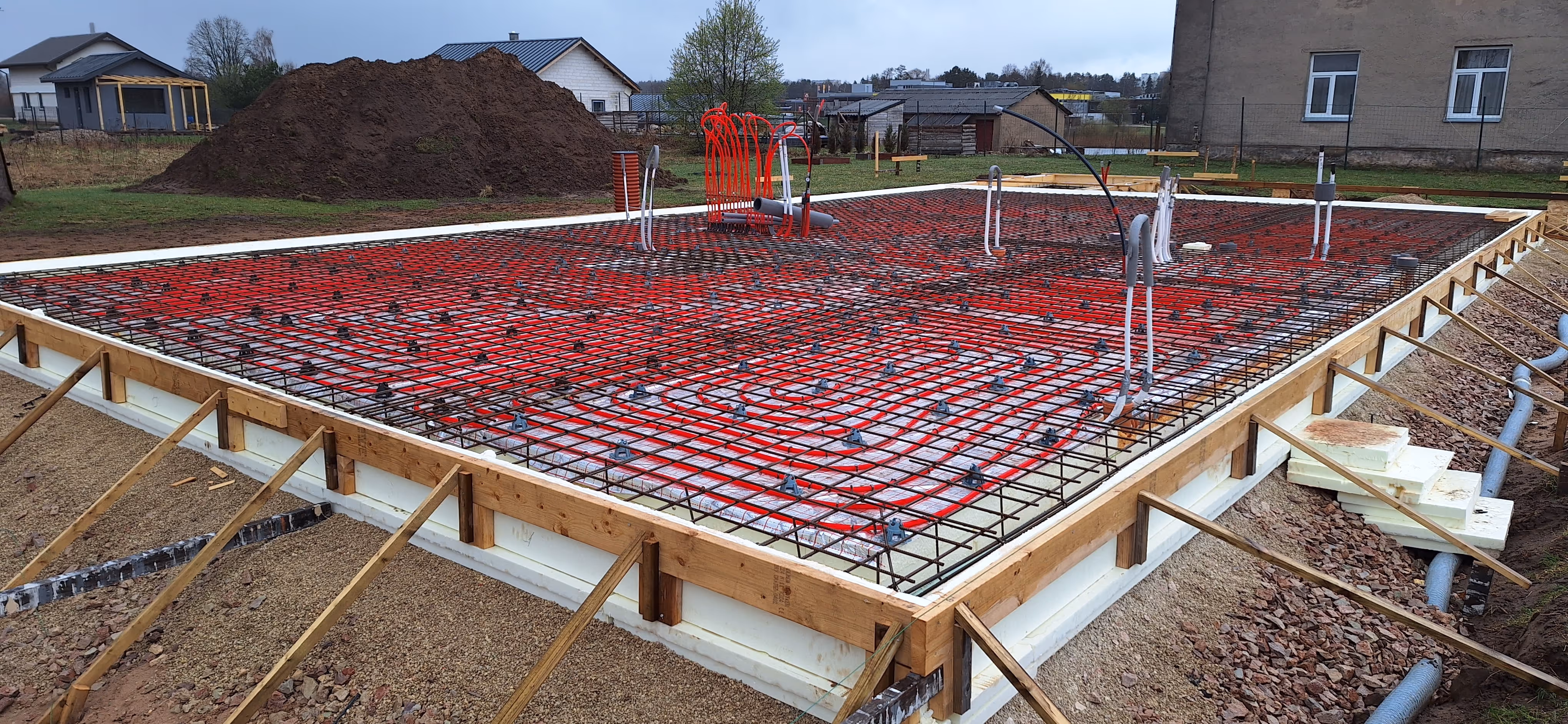 Construction site showing a wooden-framed foundation with red and black piping and metal rebar grid for underfloor heating installation.