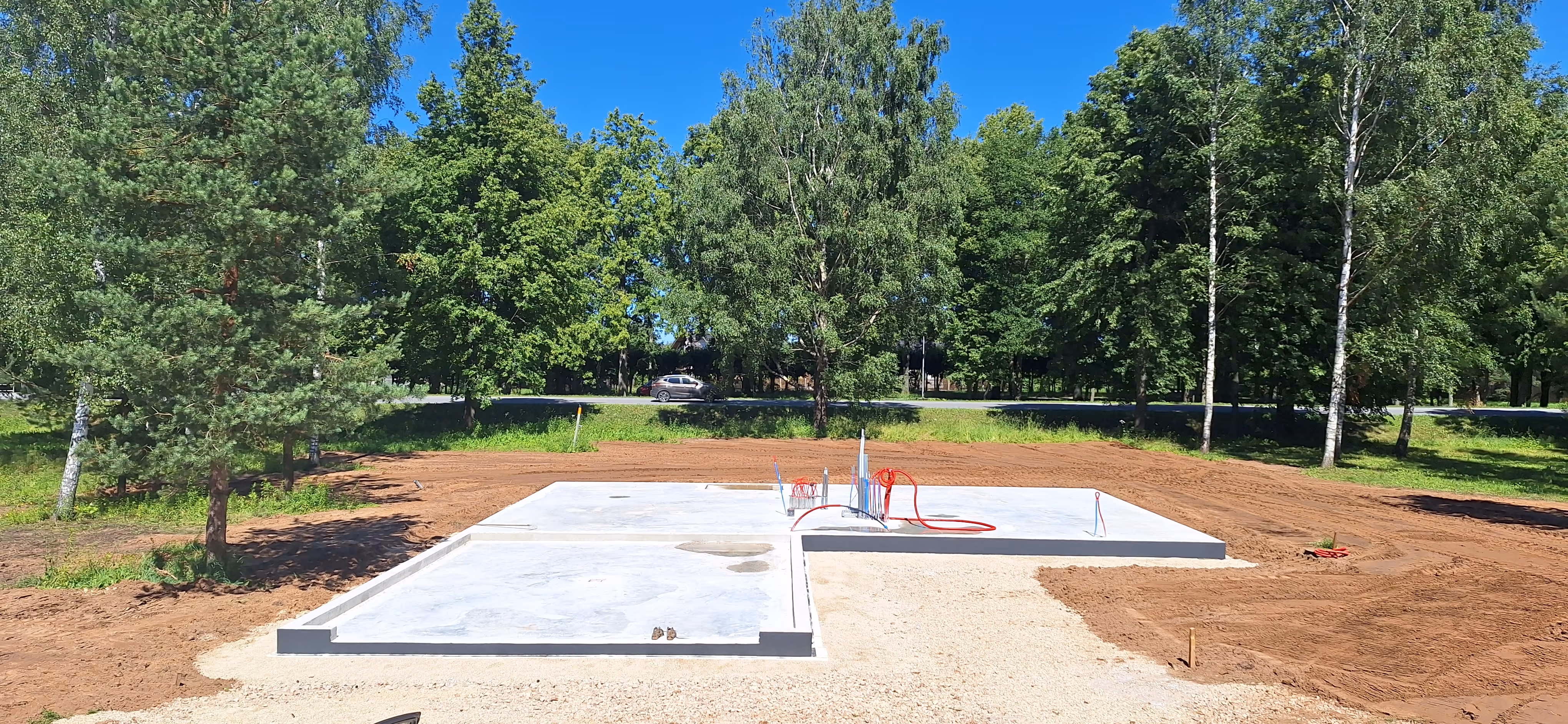 Concrete foundation slab for a building under construction in a clearing surrounded by green trees and a clear blue sky.
