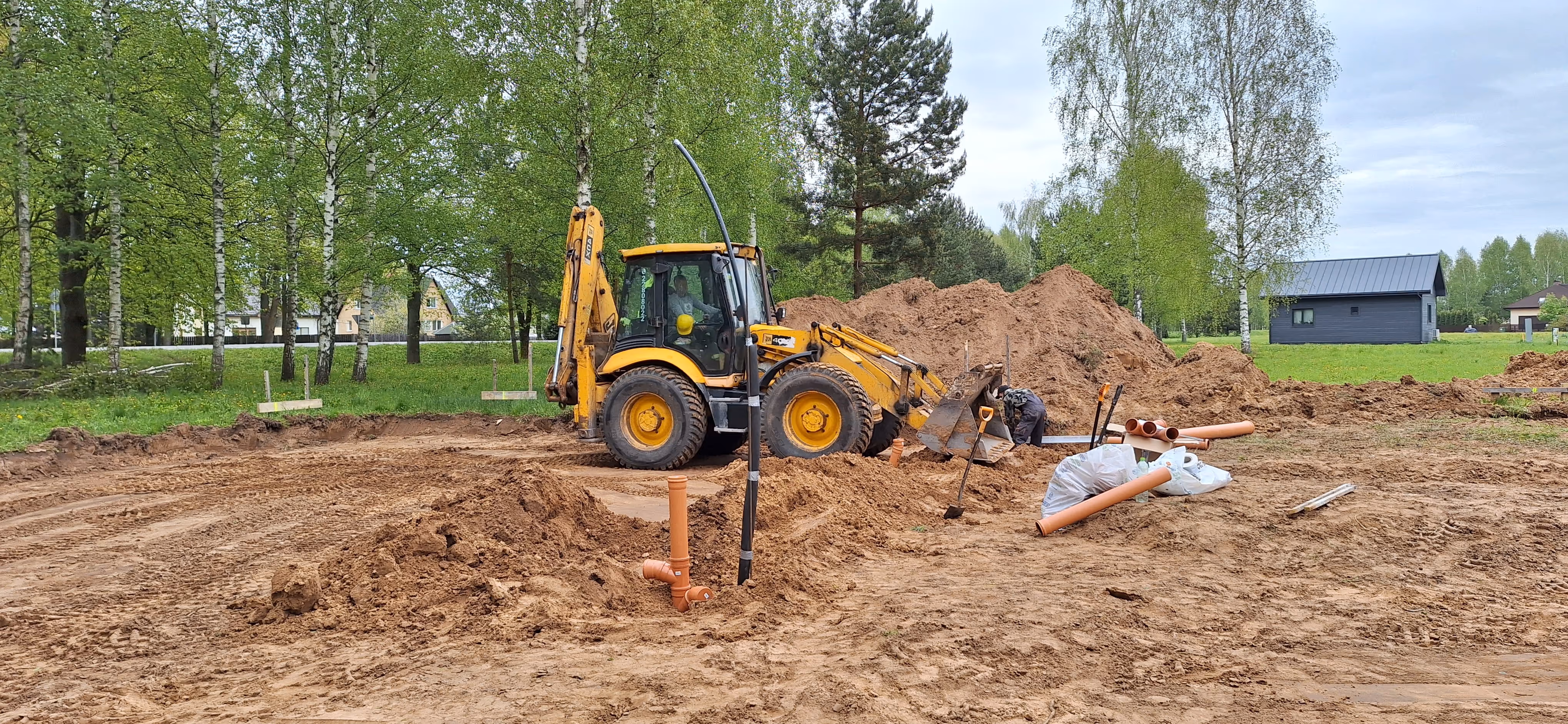 Yellow backhoe loader at a construction site with worker handling pipes and dirt piles in a green park area.