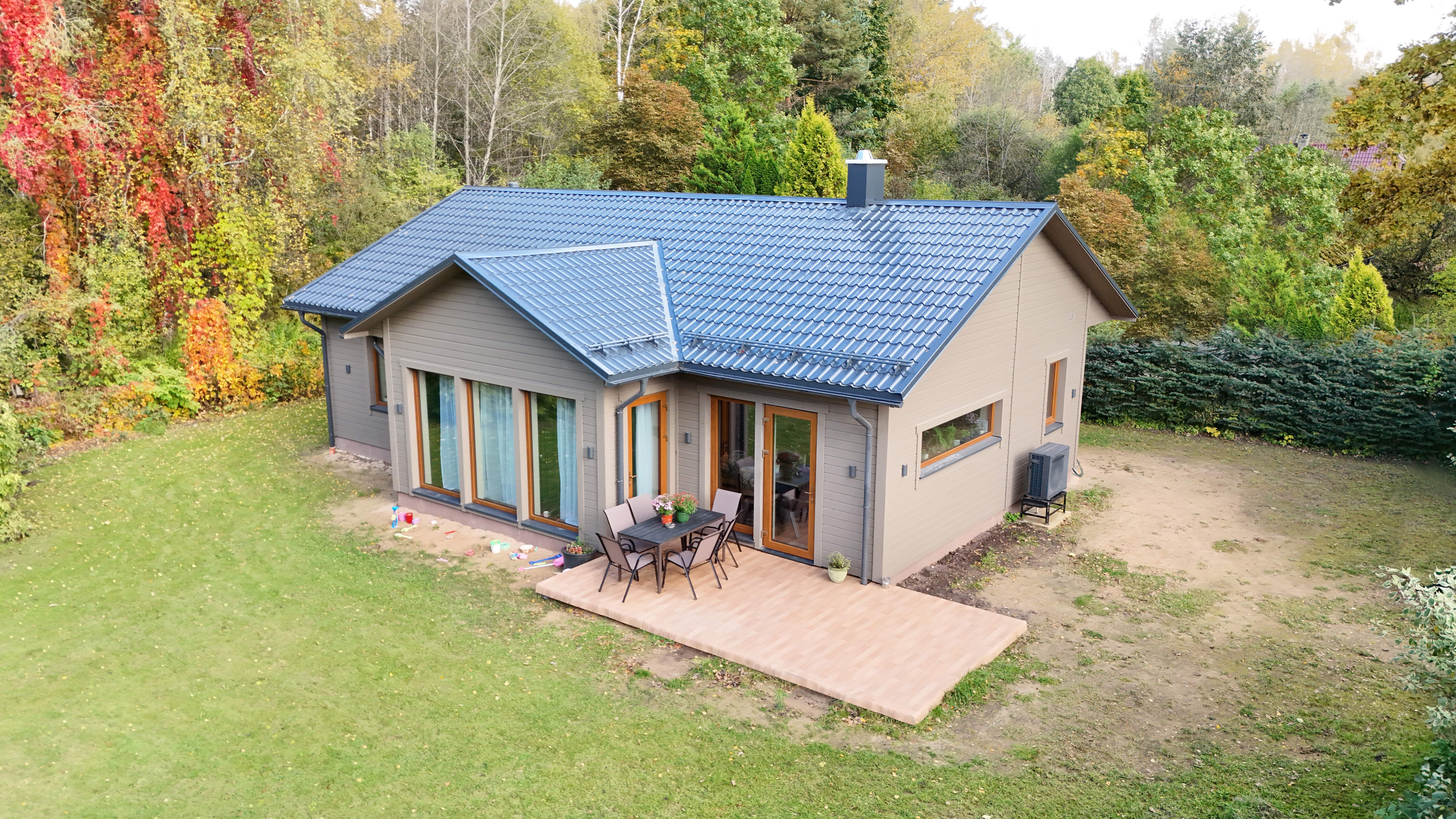 Modern single-story house with a blue metal roof and beige siding, surrounded by a green lawn and colorful autumn trees.