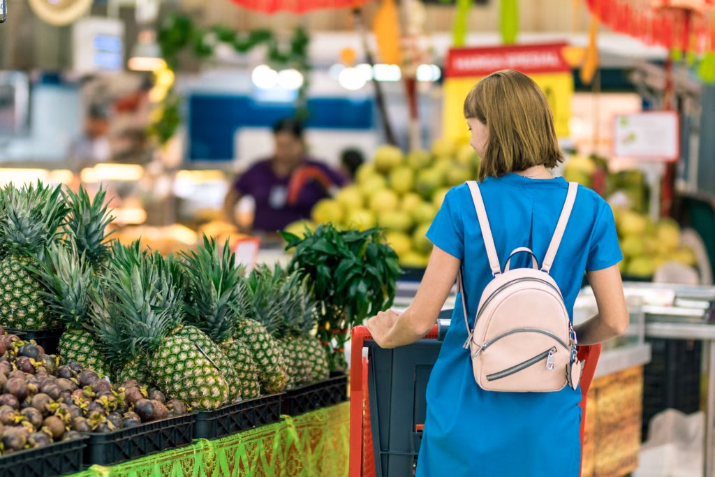 Woman in a blue dress shopping at a fruit stand with pineapples and mangosteen.
