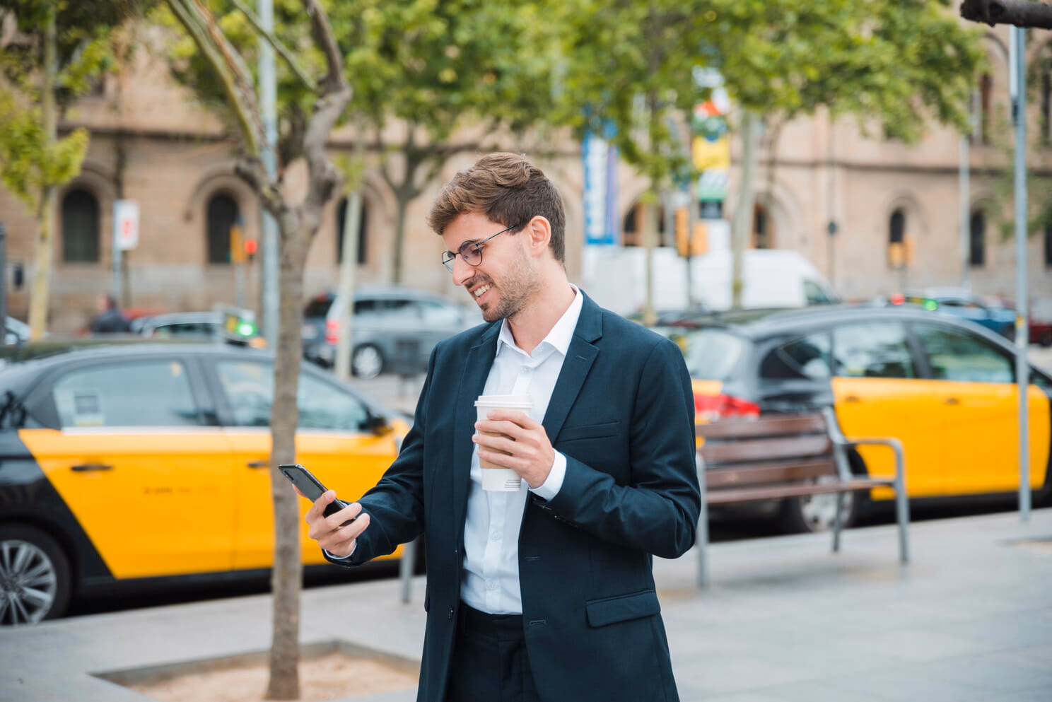 Man in a suit and glasses holds coffee and looks at phone on a city street.