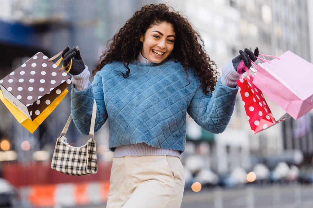 Smiling woman with curly hair holds shopping bags on a city street.