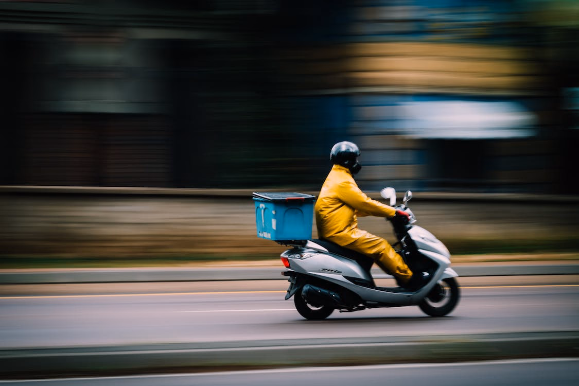 Food delivery rider in yellow suit speeds by on gray moped with a blue delivery box.