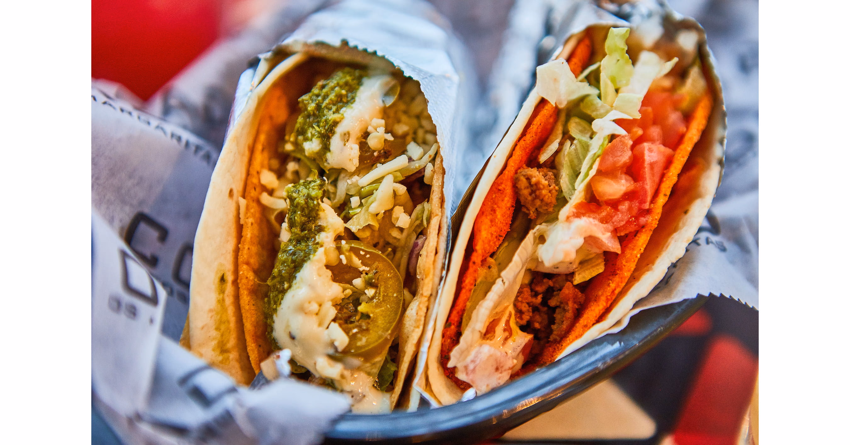 Two tacos are in a basket; one with green sauce and slaw, one with red shell, ground meat, lettuce, and tomato.