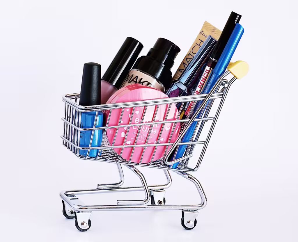Miniature shopping cart filled with various makeup items on a white background.
