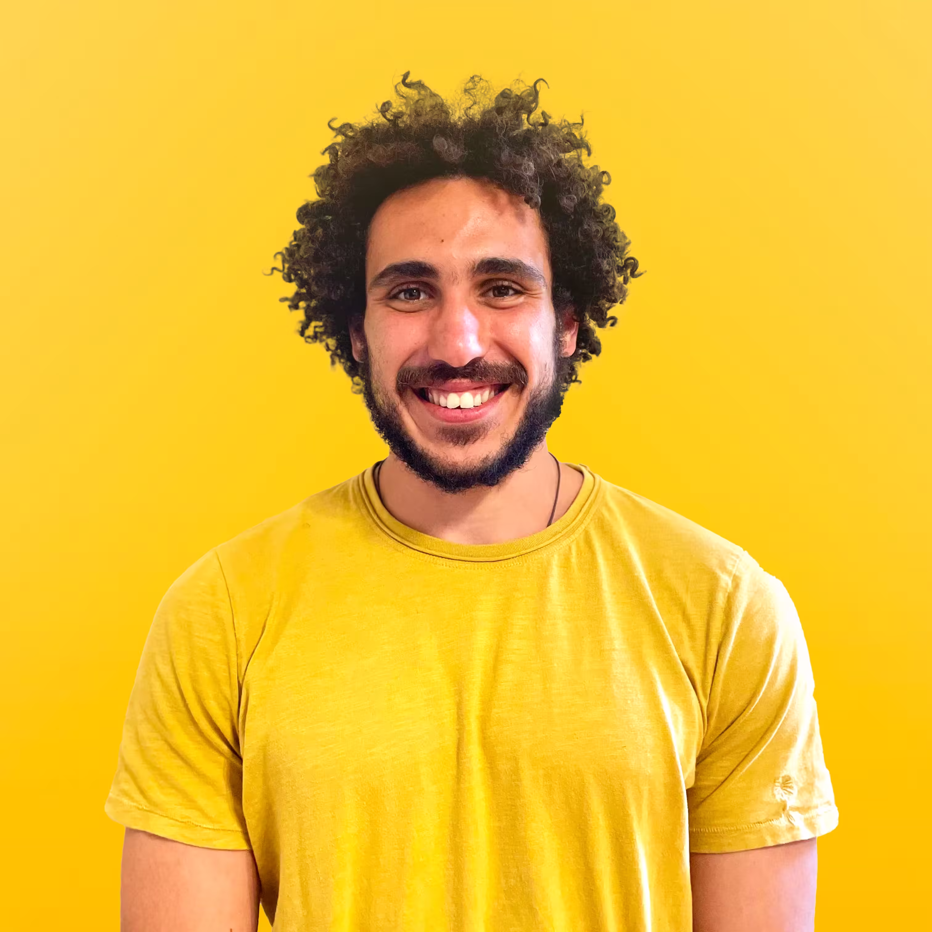 Smiling man with curly hair and a beard wearing a yellow t-shirt against a yellow background.