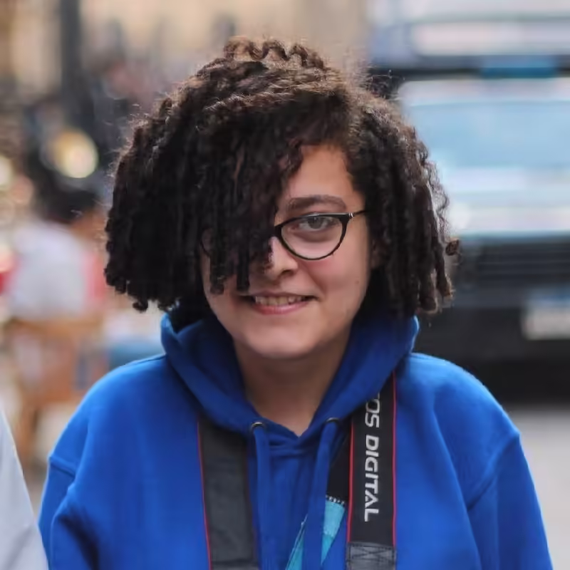 Person with dark curly hair partly obscuring their face, wearing glasses and a blue hoodie, with a camera strap.