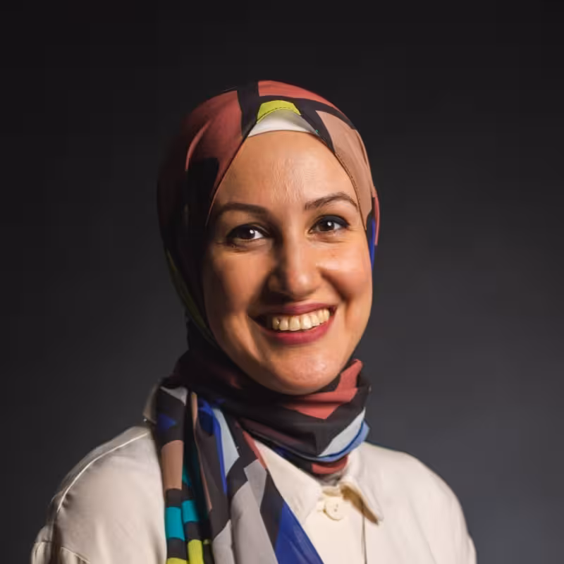 Woman with colorful headscarf smiles against dark gray background.