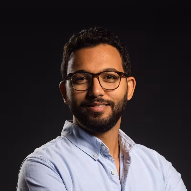 Man with dark hair, glasses, and beard is wearing a blue button-down shirt and smiling.