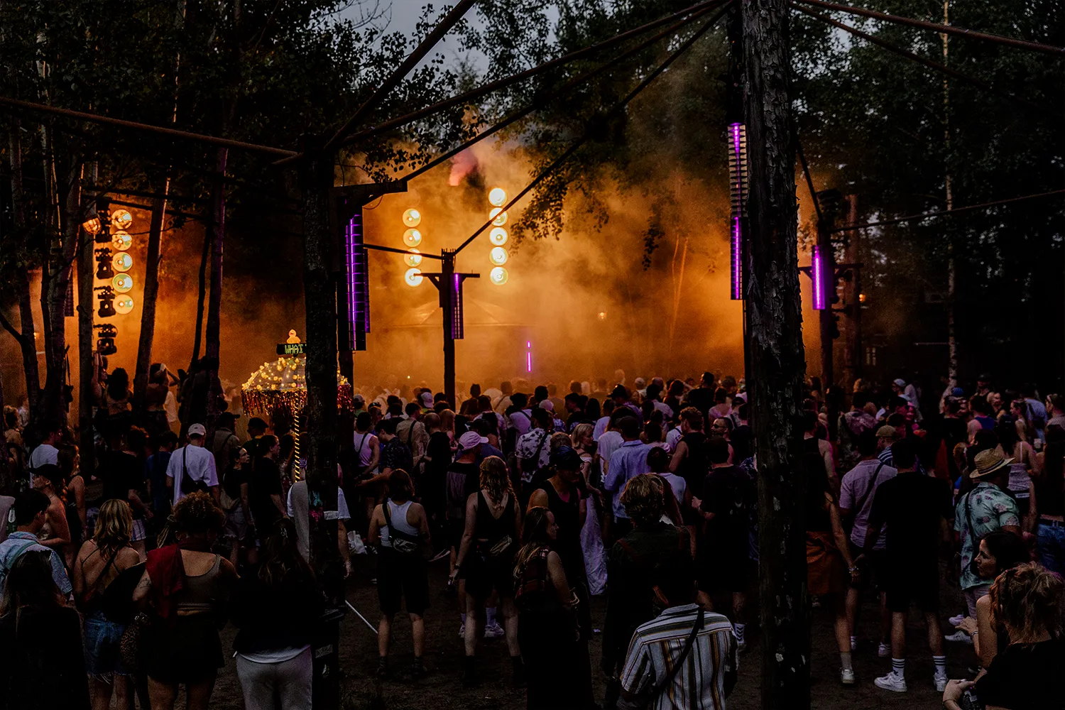 Eine Menge an Menschen im Wald, im Hintergrund ist eine Bühne im dichten Nebel zu sehen.