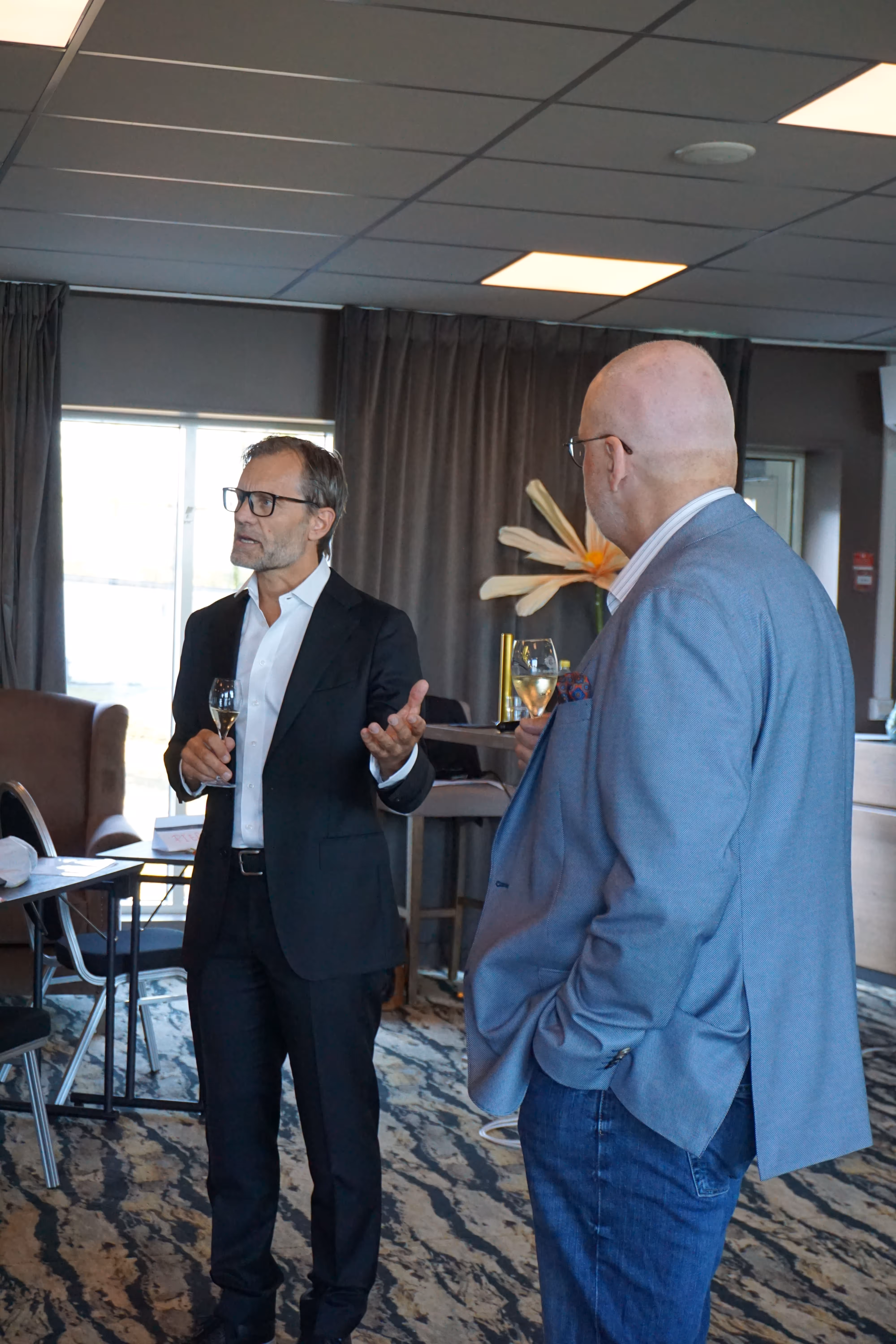 Två män i ett rum, en i svart kostym och glasögon håller ett glas vin och gestikulerar medan den andra i blå kavaj och jeans lyssnar.