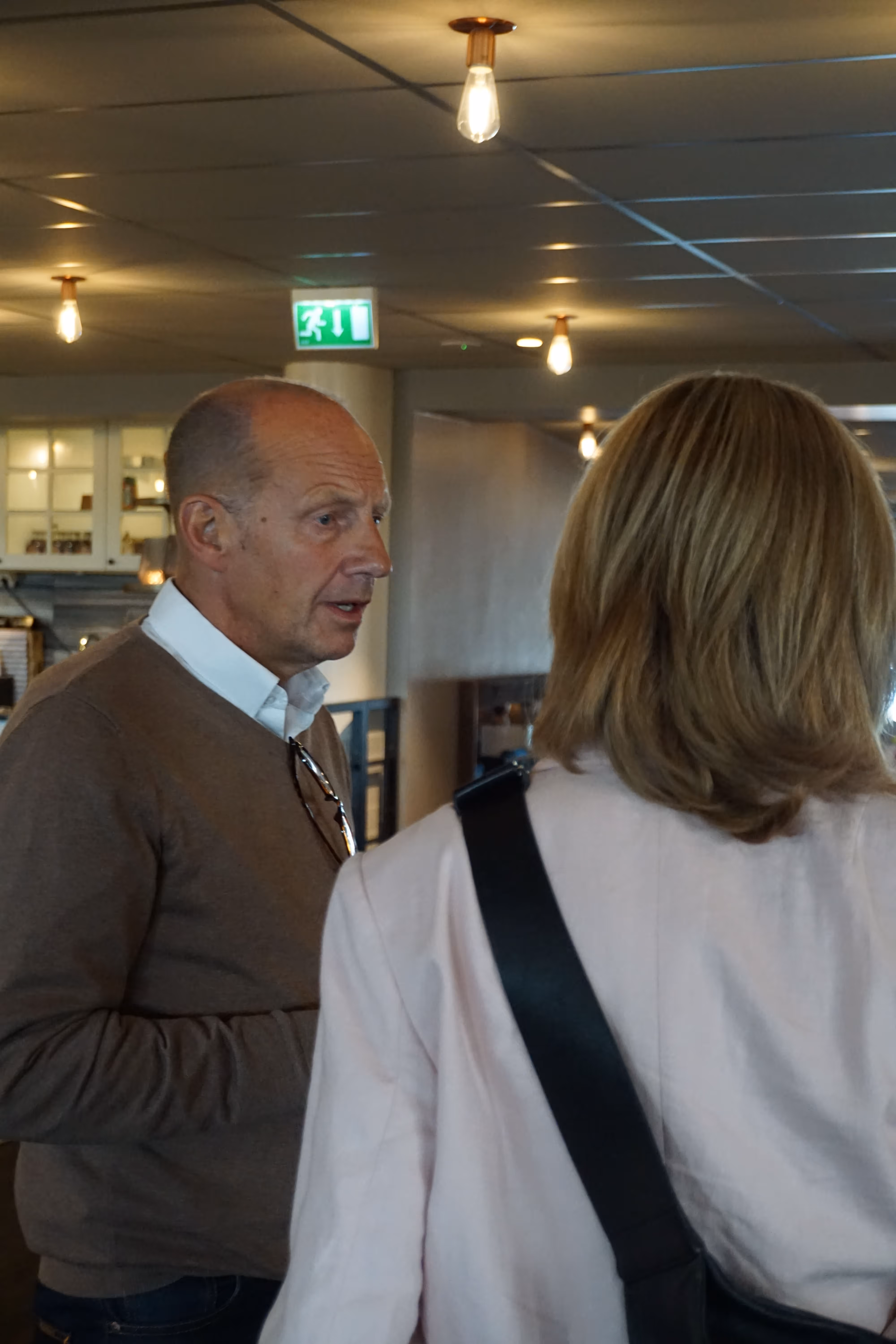 En man i brunt plagg samtalar med en kvinna med ljust hår och ljus jacka i ett väl upplyst inomhusrum.