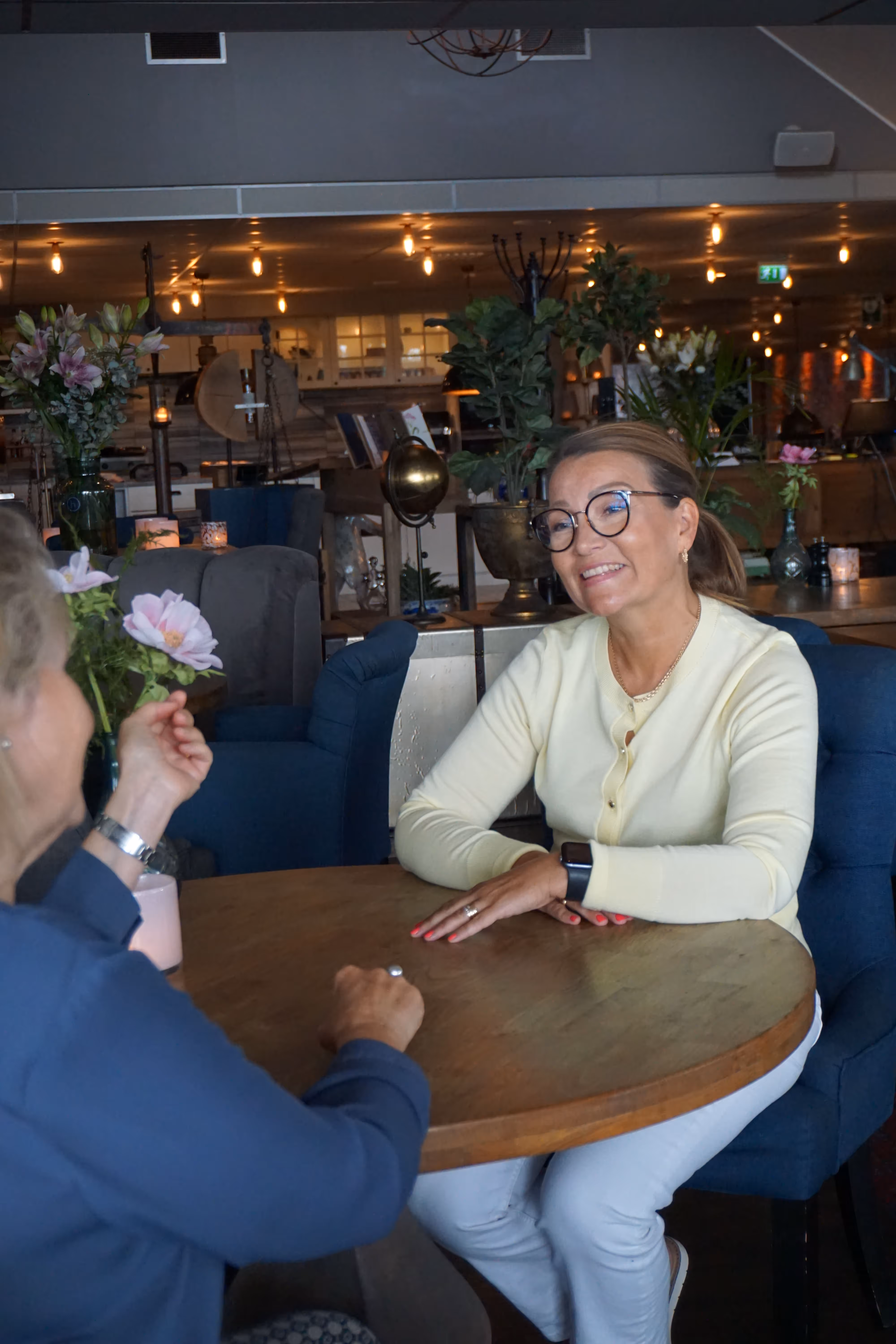 Två kvinnor sitter vid ett runt bord i ett mysigt rum och pratar medan den ena ler.