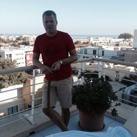 Man i röd t-shirt och beige shorts lutar sig mot ett räcke på en balkong med utsikt över en stad och havet i bakgrunden.