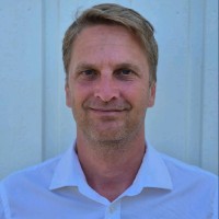 Porträtt av en leende man med kort blont hår i en vit skjorta framför en ljus bakgrund.