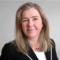 Porträtt av en leende kvinna med långt ljust hår i svart kavaj mot grå bakgrund.