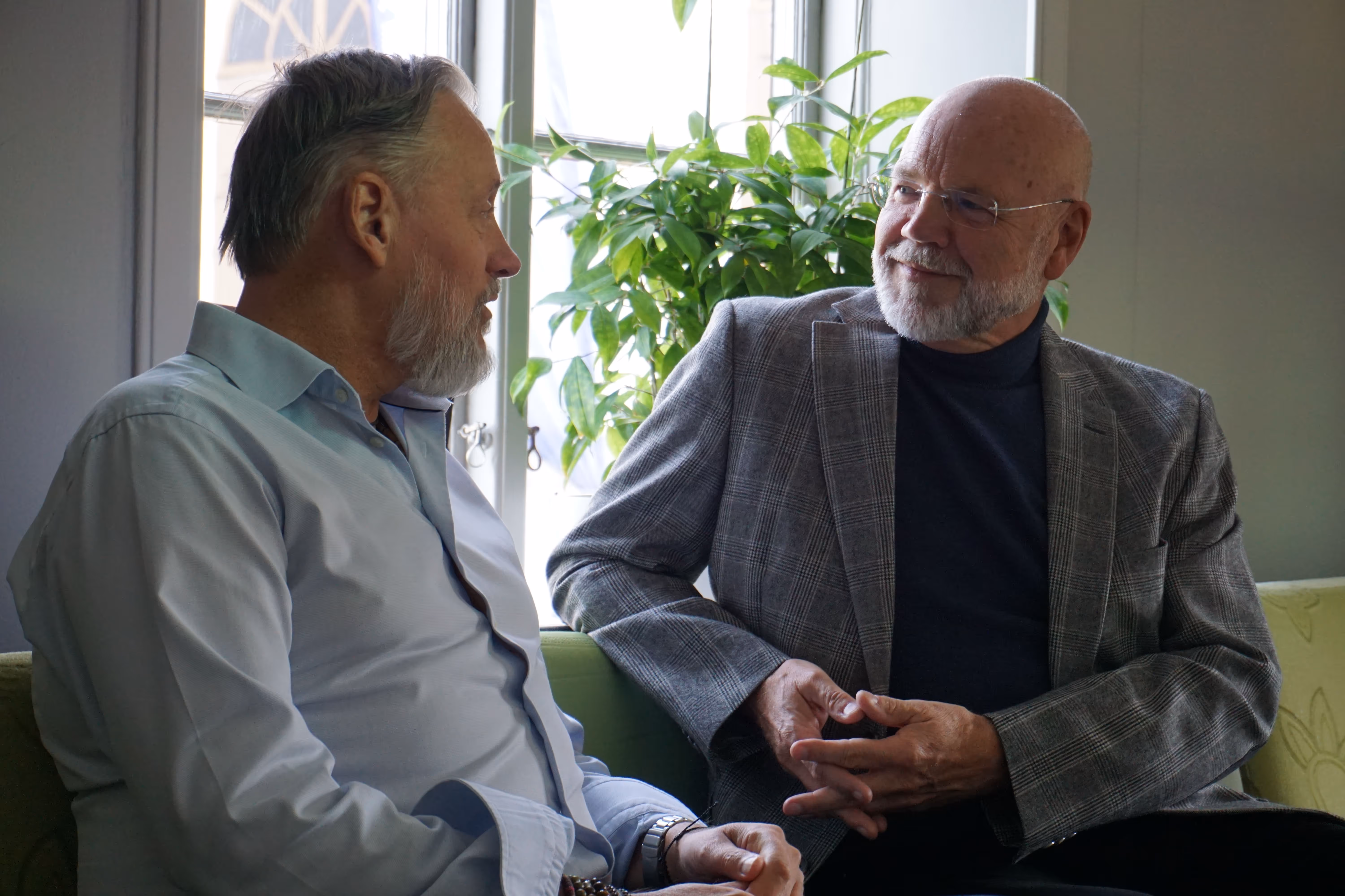 Två äldre män med skägg sitter och samtalar avslappnat i en soffhörna vid ett fönster med en stor grön växt i bakgrunden.