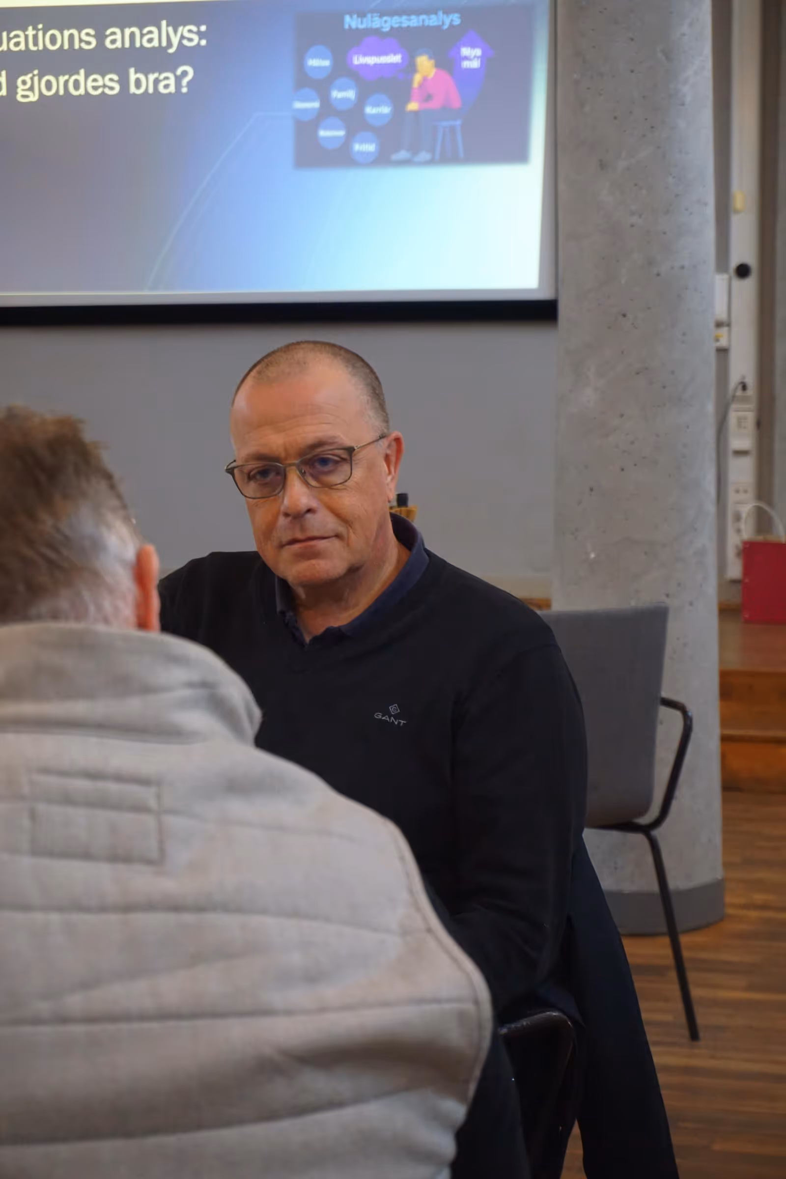 En man med glasögon sitter och lyssnar uppmärksamt mot en annan person under ett möte med en presentationsskärm i bakgrunden.