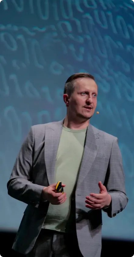 Man wearing a gray blazer and green shirt speaking on stage with a headset microphone and abstract blue background.
