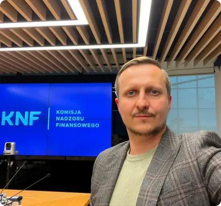 Man with short blonde hair and light facial hair wearing a gray checkered blazer and light green sweater, sitting in a conference room with a wooden ceiling and a screen displaying 'KNF Komisja Nadzoru Finansowego'.