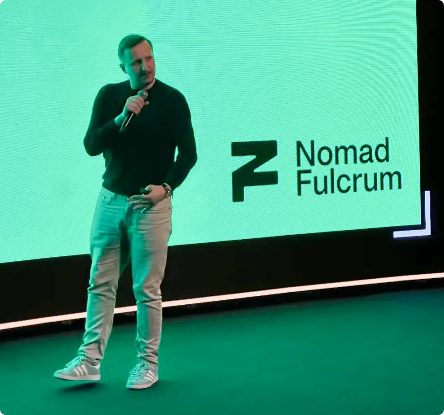 A man holding a microphone stands on stage in front of a screen displaying the Nomad Fulcrum logo.