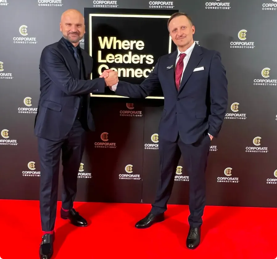 Two men in suits shaking hands on a red carpet in front of a backdrop with Corporate Connections logos and a sign reading 'Where Leaders Connect'.