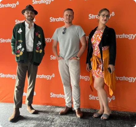 Three adults standing in front of an orange backdrop with repeated 'Strategy' logos: a man in a hat and patterned cardigan, a man in a gray t-shirt and light pants, and a woman in a colorful dress with a black cardigan.
