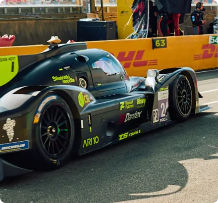 Black prototype race car with yellow and green decals parked on a race track near a yellow DHL barrier.