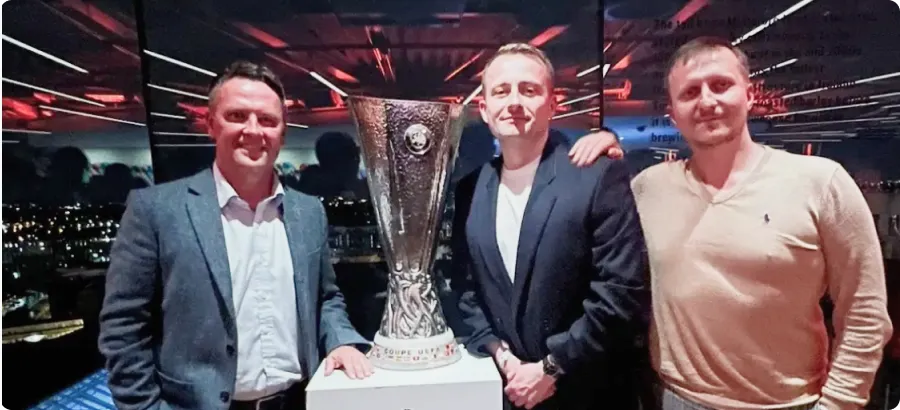 Three men standing indoors around a tall UEFA Europa League trophy on a white pedestal with city lights visible through the window behind them.