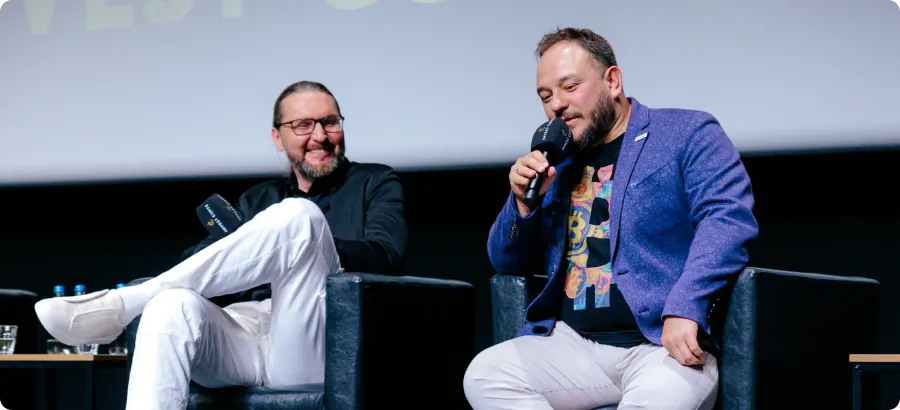 Two men sitting in black armchairs on a stage, one speaking into a microphone while the other listens and smiles.