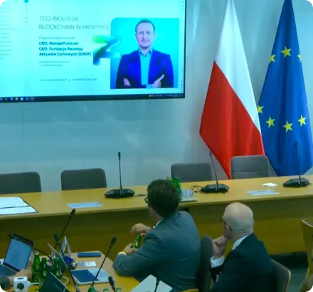 Two men in a conference room looking at a presentation on blockchain technology displayed on a screen, with Polish and European Union flags beside it.