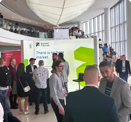 People networking inside a bright convention center near a Nomad Fulcrum booth with a message saying 'There is no bear market.'