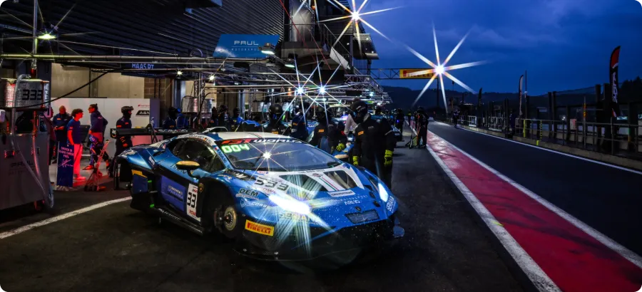 Blue race car with headlights on in a nighttime pit stop with crew members and illuminated garage area.