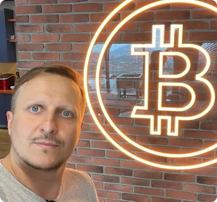 Man with short hair and a beard taking a selfie in front of a brick wall with a large illuminated Bitcoin symbol.