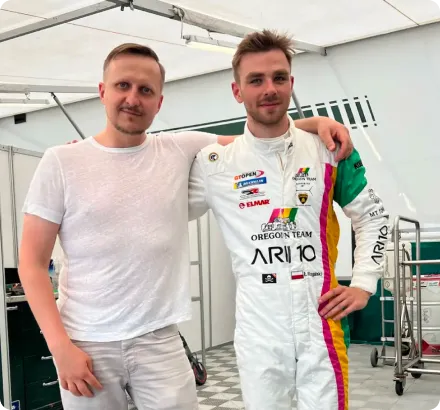 Two men standing side by side under a tent, one in a white T-shirt and shorts, the other in a white racing suit with colorful stripes and sponsor logos.