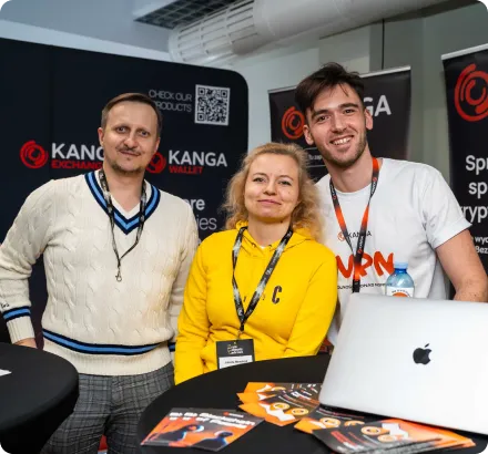 Three people smiling at a promotional booth with Kanga Exchange and VPN branding, standing behind a table with brochures and a laptop.